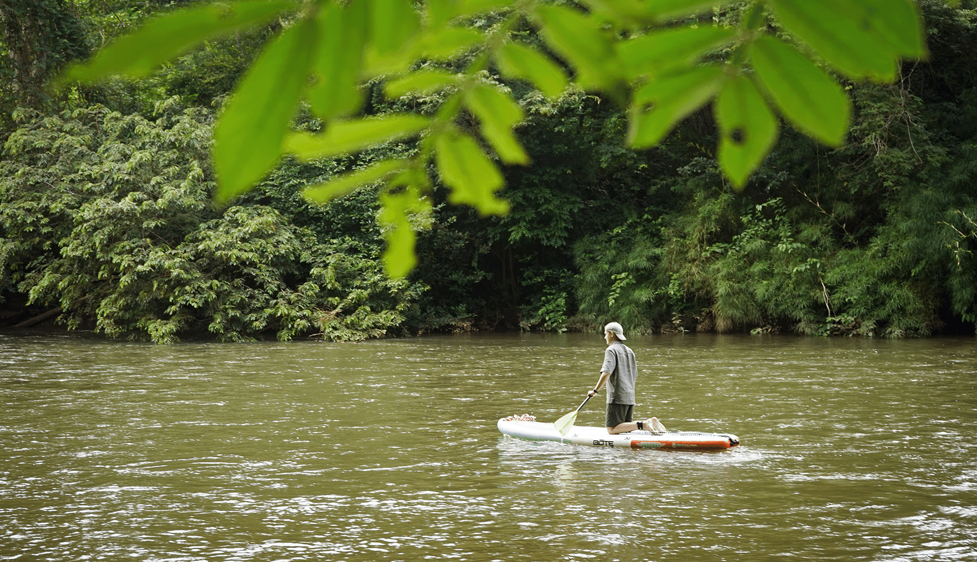 A person paddles a small boat on a calm river surrounded by lush greenery.