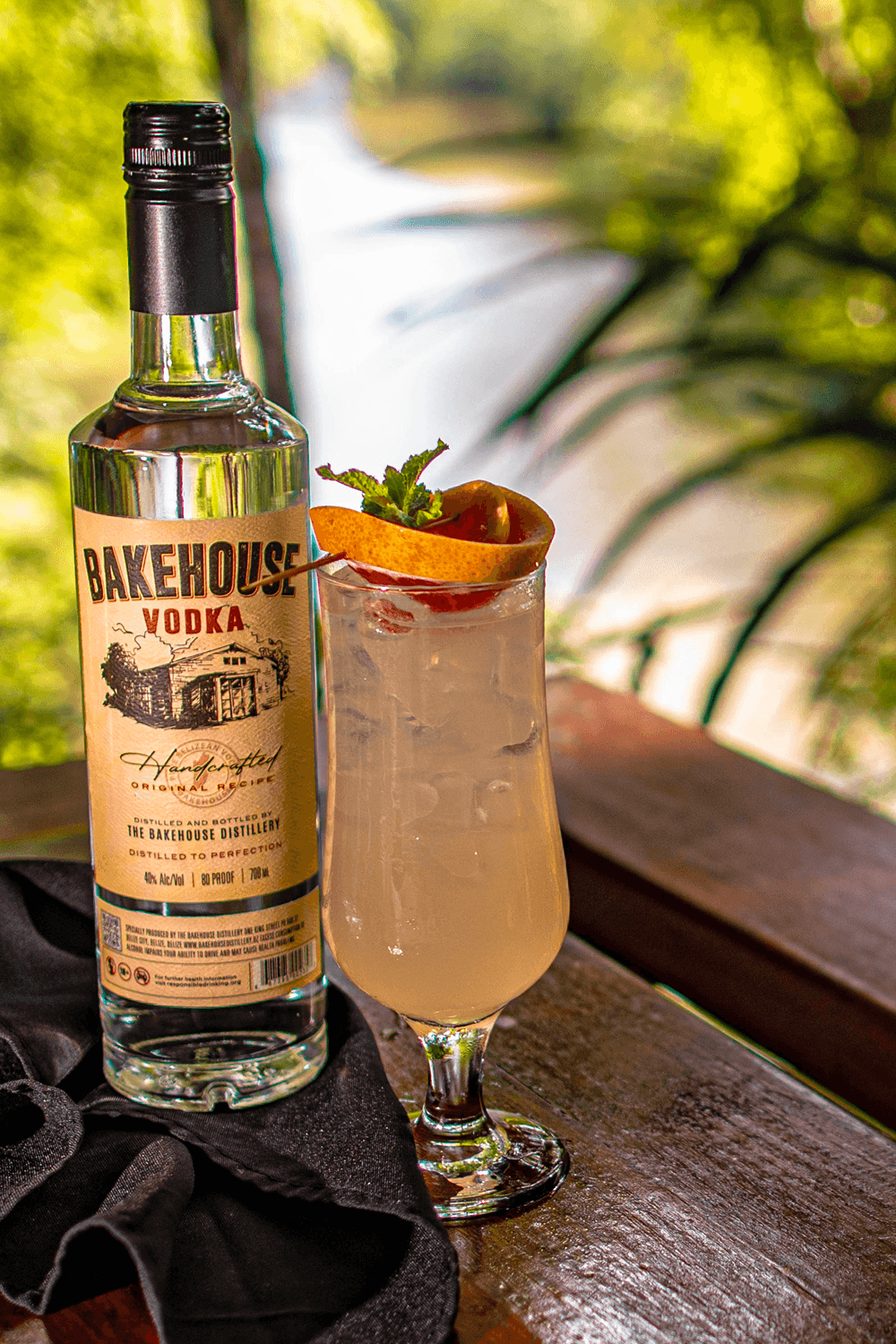 A bottle of Bakehouse Vodka next to a cocktail garnished with a slice of pear and mint, set against a lush, blurred background.