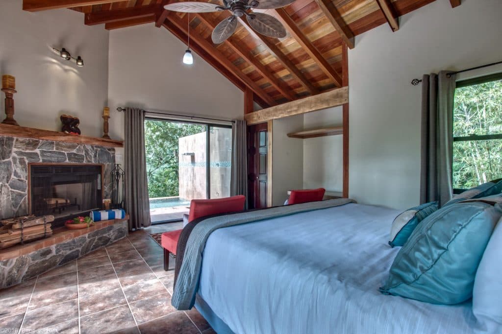 Cozy bedroom with a stone fireplace, wooden ceiling, and large windows leading to an outdoor area.
