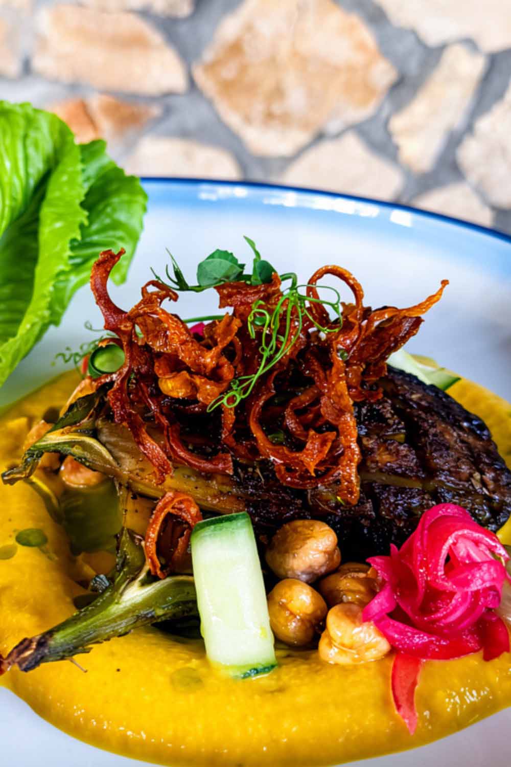 A vibrant dish featuring grilled protein topped with crispy onions, served on a bed of smooth purée and garnished with vegetables and herbs.