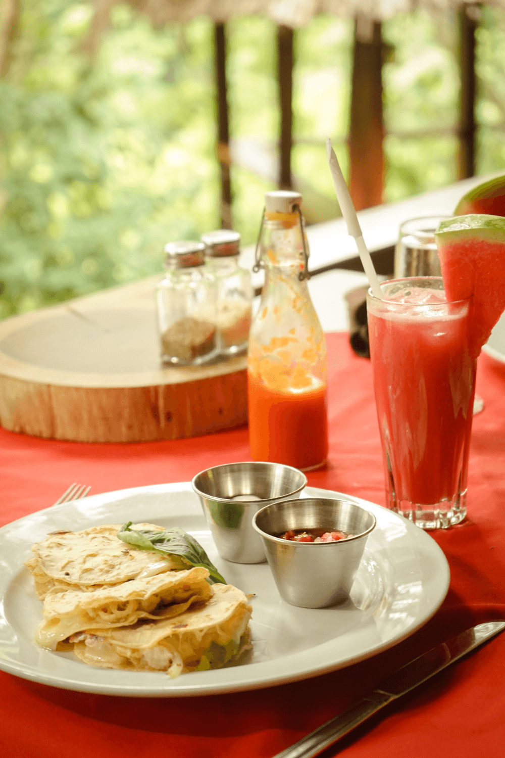 A plate of tortillas with fillings, two small bowls of sauce, and a glass of watermelon juice on a red tablecloth.