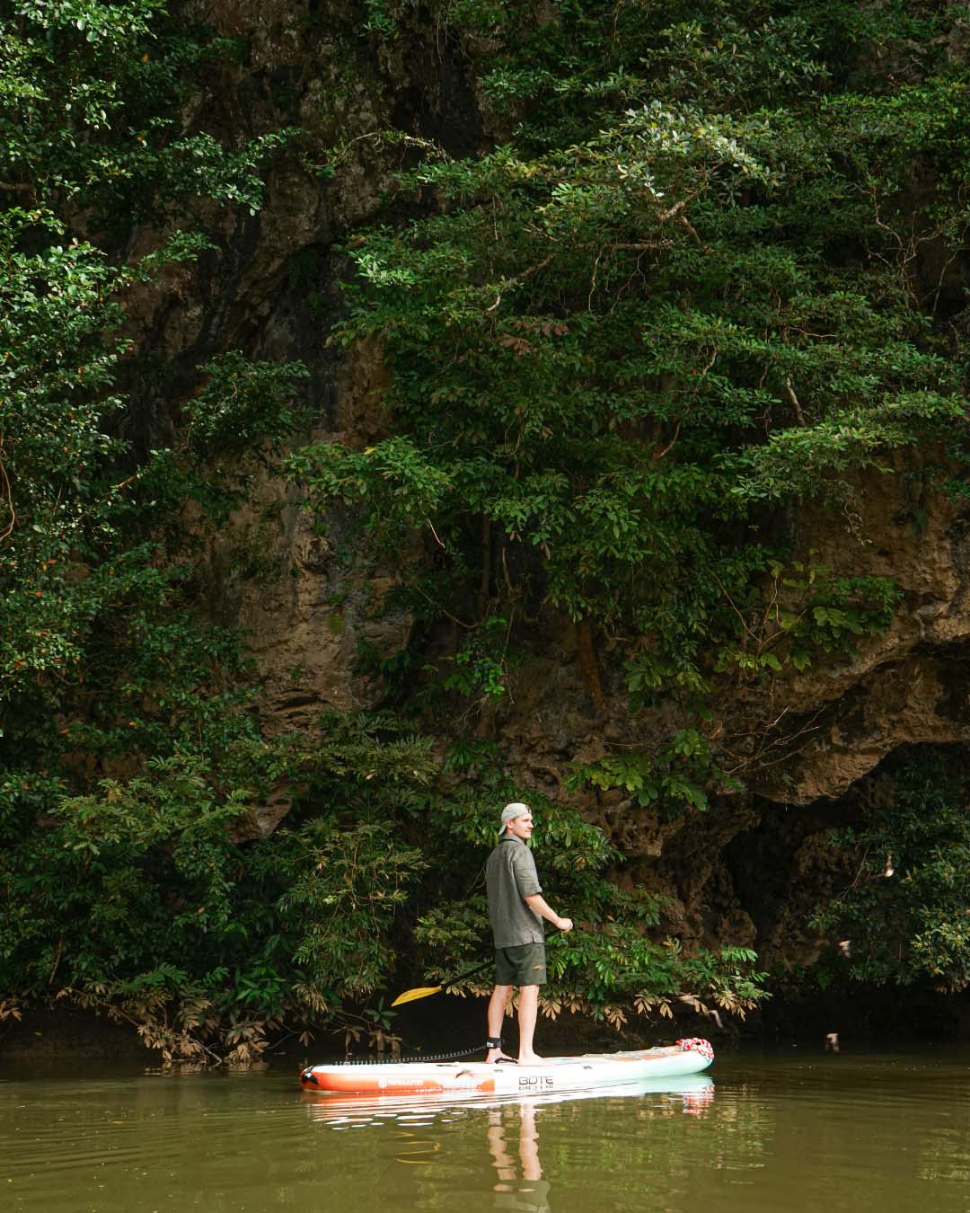 A person stands on a paddleboard in a lush, green river surrounded by cliffs.