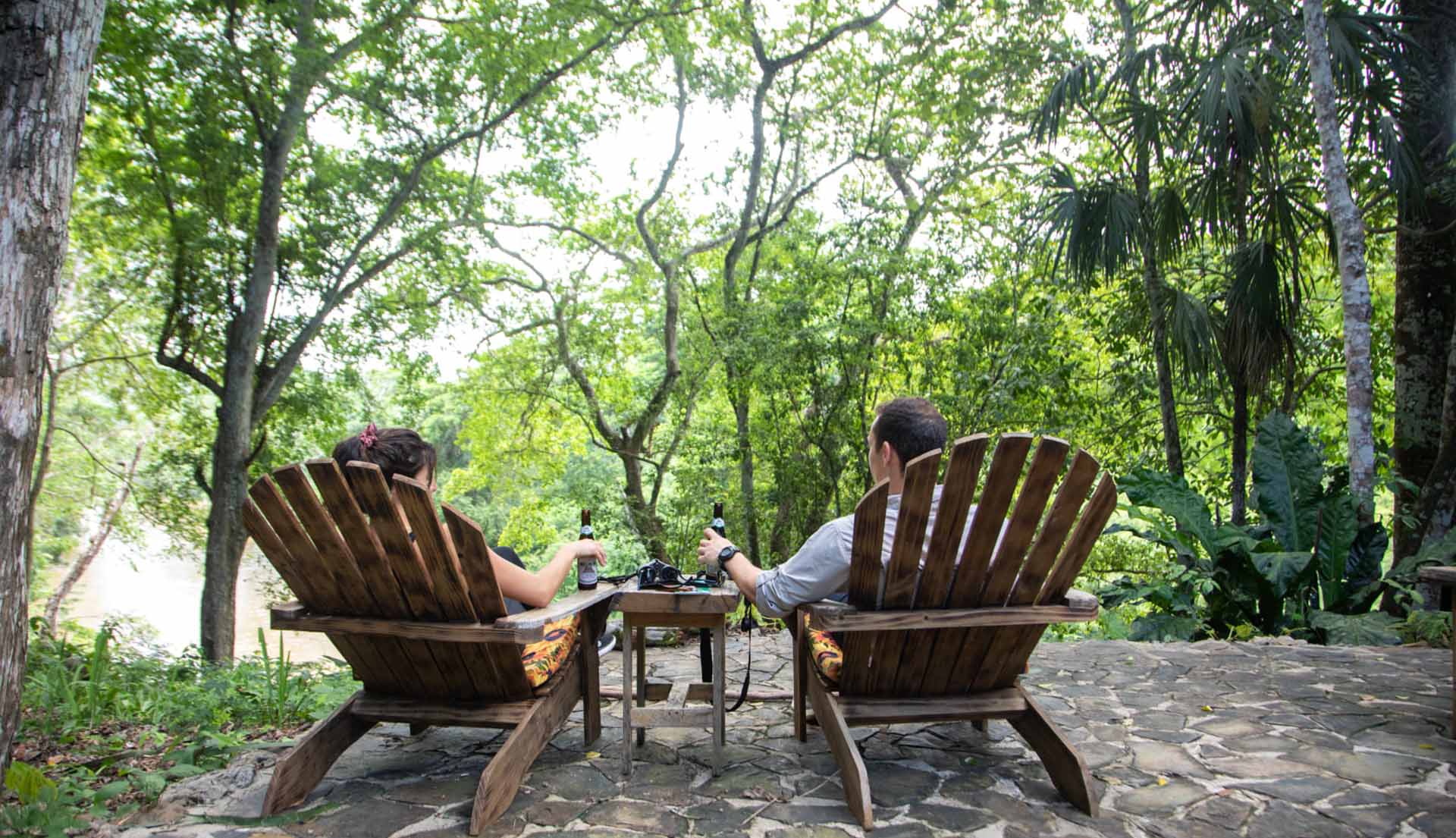 A couple relaxes in wooden chairs, enjoying drinks amidst lush greenery.