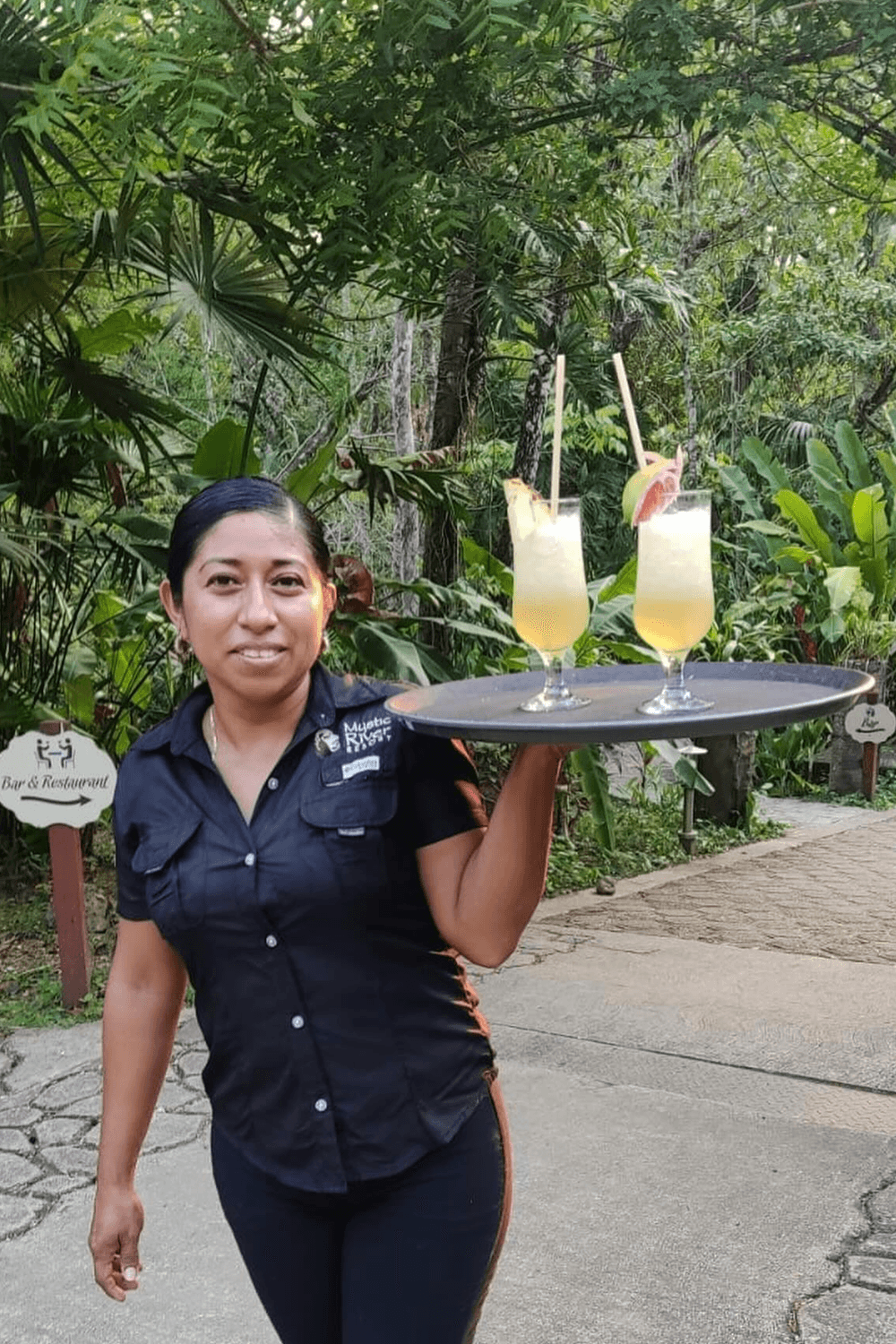 A server carries a tray with two cocktails in a lush, tropical setting.