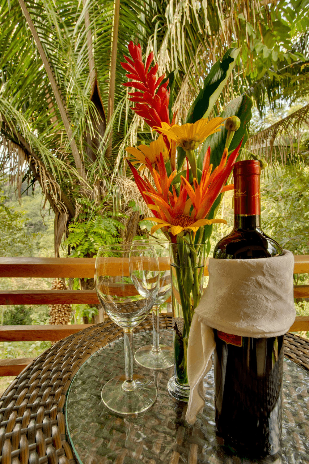 A wine bottle wrapped in a napkin sits beside two empty glasses and a vase of colorful tropical flowers on a glass table against a lush green background.