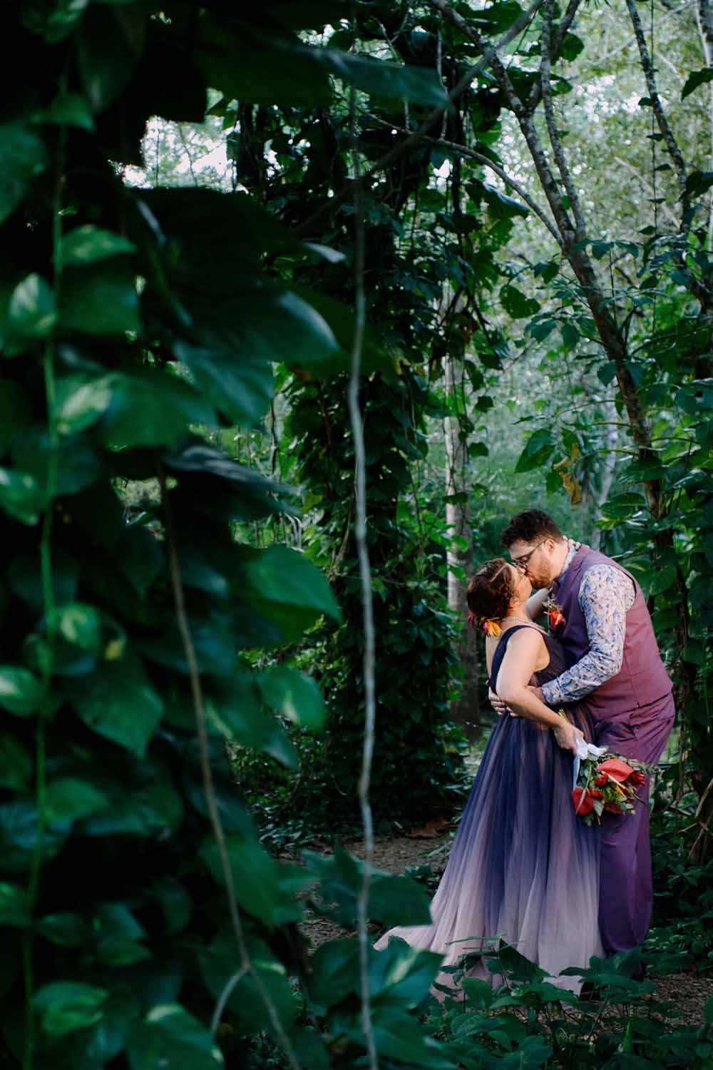 A couple embraces and shares a kiss surrounded by lush greenery.