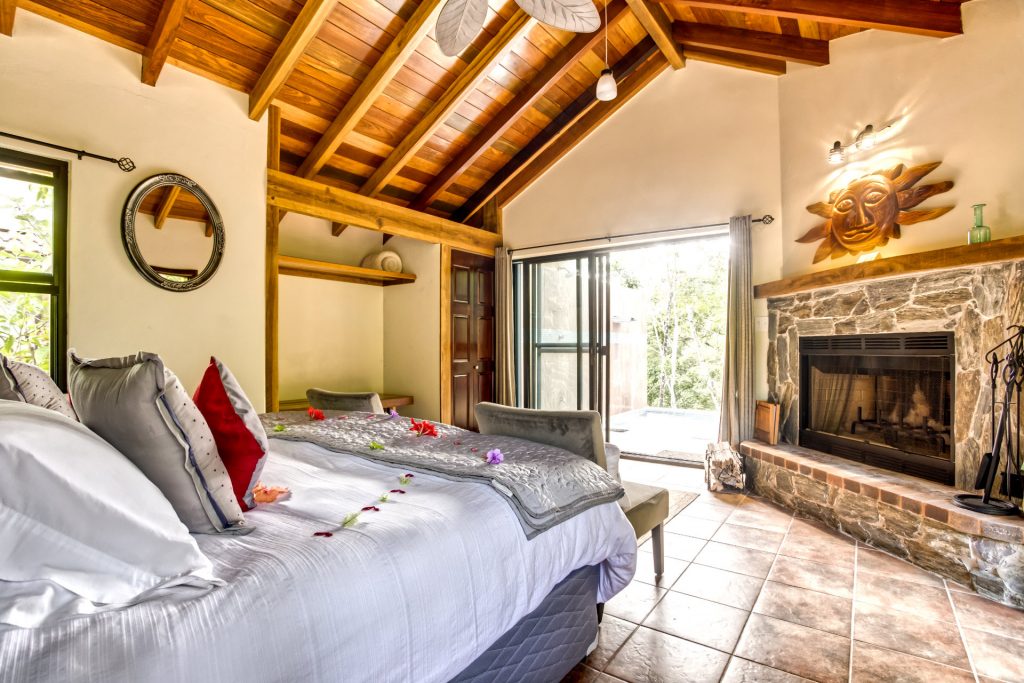 Cozy bedroom with a wooden ceiling, stone fireplace, and large windows opening to a scenic view.