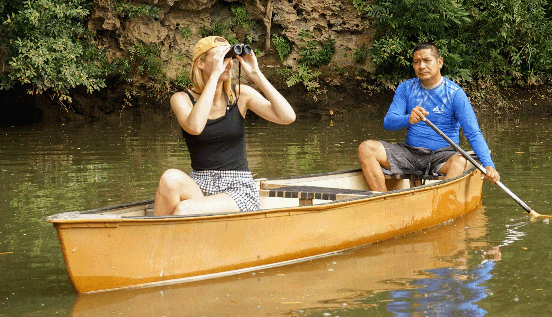 A woman in a canoe uses binoculars while a man paddles alongside her in a calm waterway surrounded by greenery.
