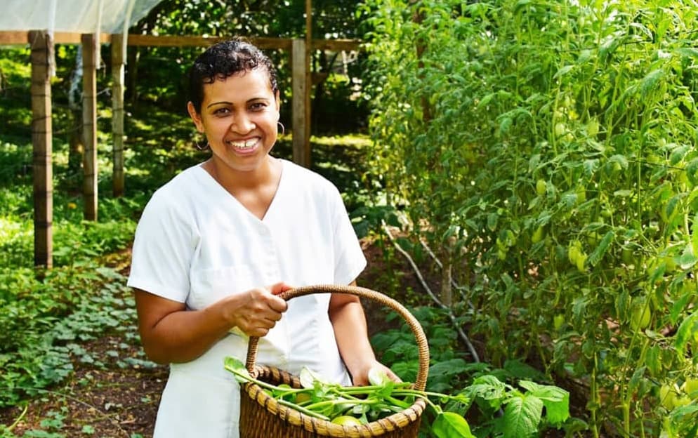 A woman in a white dress smiles while holding a basket of greens in a lush garden.