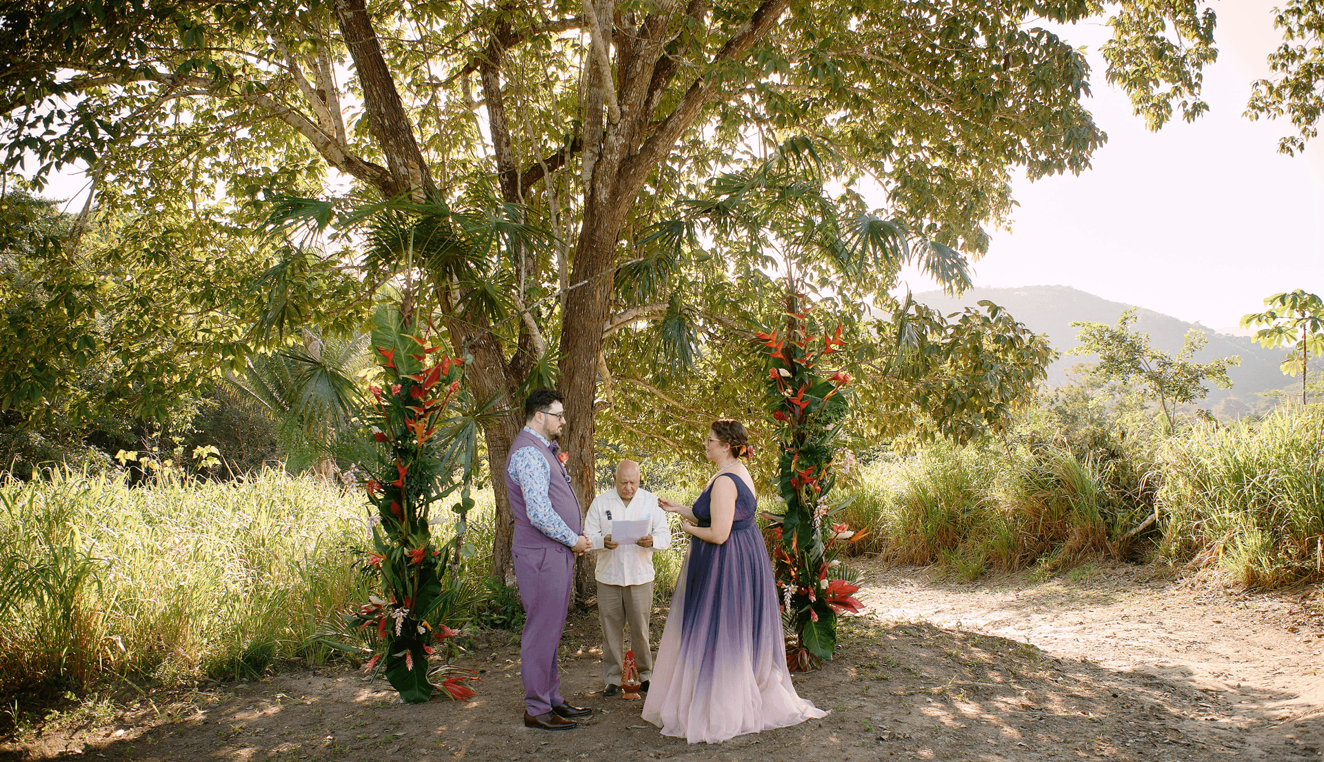 A couple stands under a large tree during a wedding ceremony, flanked by floral decorations and an officiant holding a script.