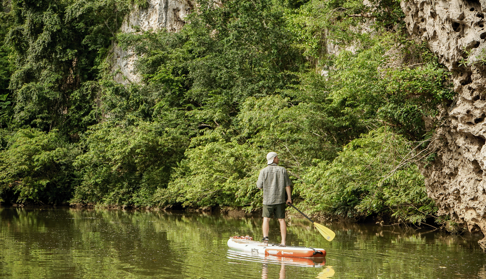 A person stands on a paddleboard in calm waters, surrounded by lush green foliage and rocky cliffs.