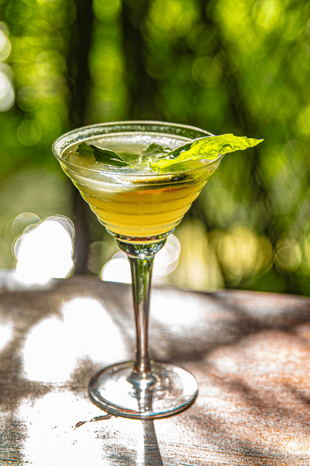 A close-up of a cocktail glass filled with a light yellow drink and garnished with fresh basil, set against a blurred green background.