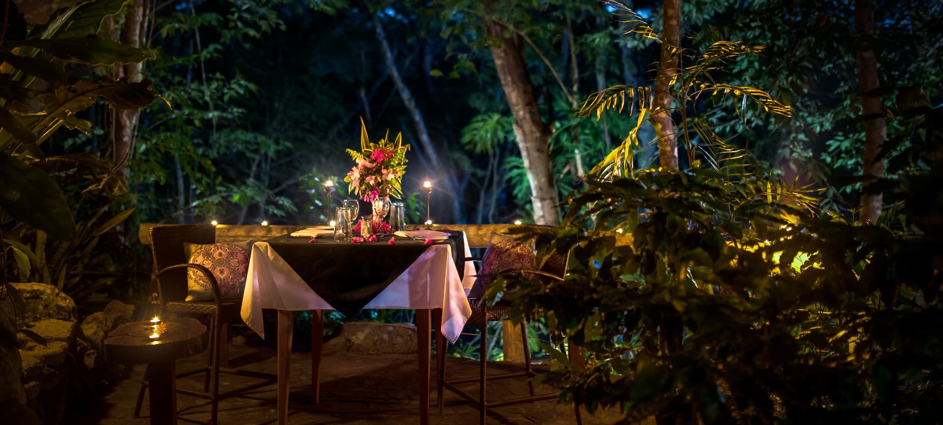 A romantic outdoor dining setup surrounded by lush greenery and soft candlelight.