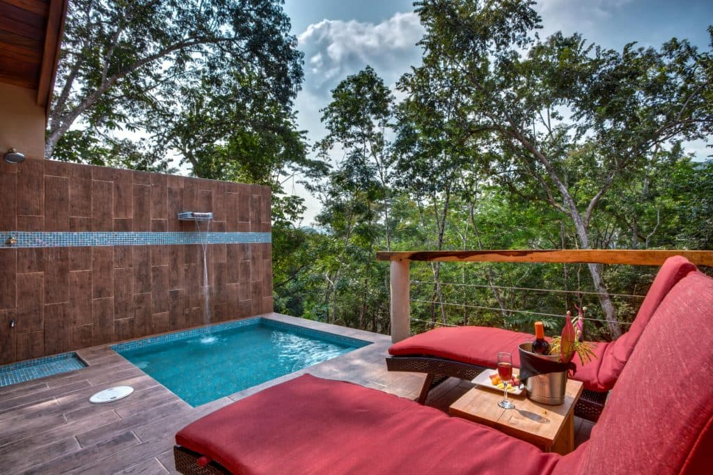 A tranquil outdoor space featuring a small pool, red lounge chairs, and a scenic view of lush greenery.