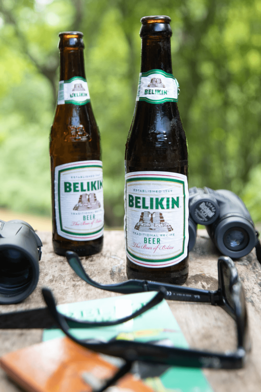 Two bottles of Belikin beer are displayed on a wooden surface alongside binoculars and sunglasses.