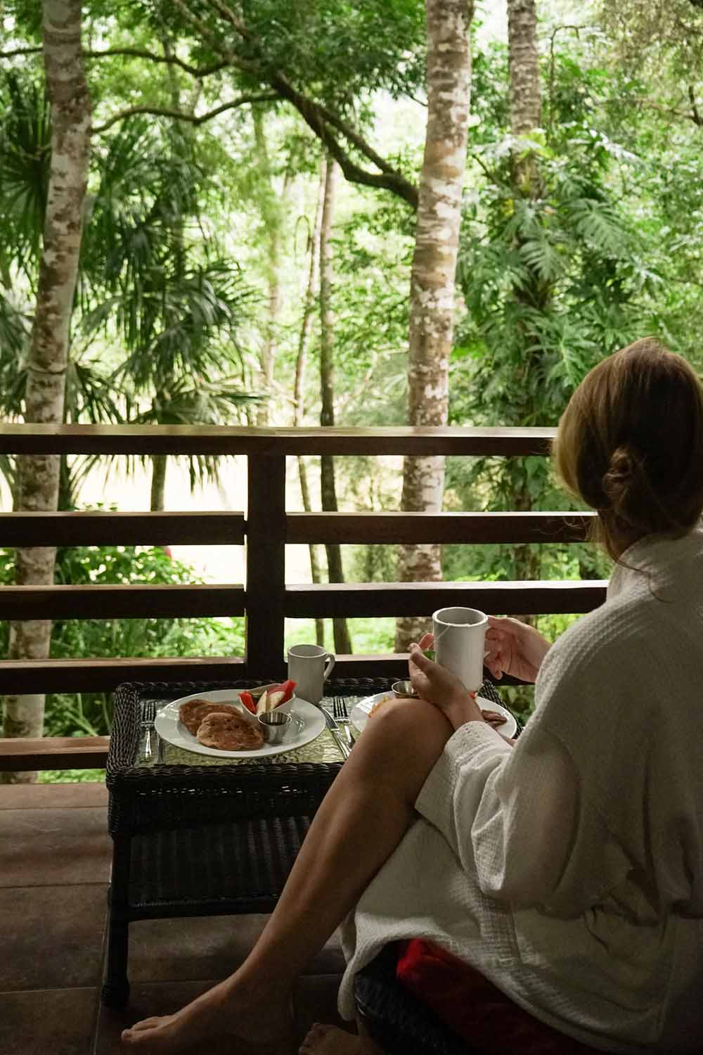 A person in a bathrobe enjoys breakfast and coffee on a balcony surrounded by lush greenery.