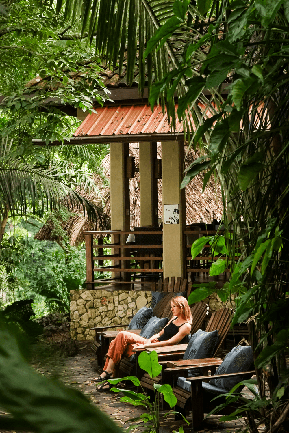 A woman relaxes on a wooden lounge chair surrounded by lush greenery.