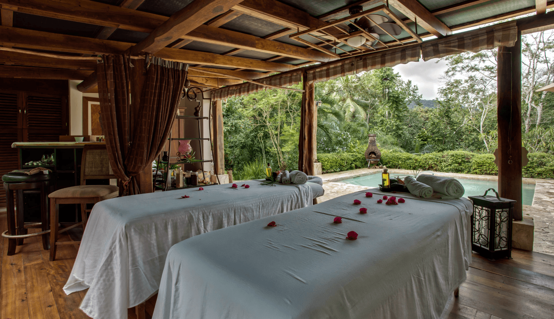 A serene spa room featuring two massage tables adorned with rose petals and a view of lush greenery.