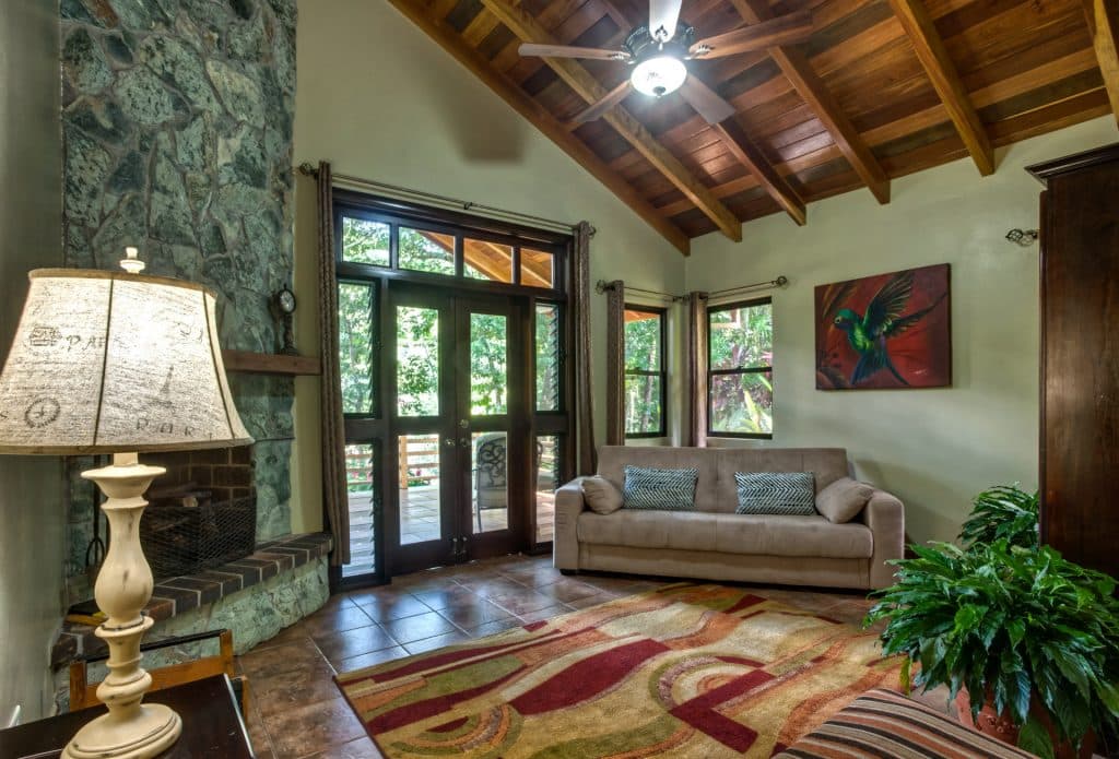A cozy living room featuring a stone fireplace, a light brown sofa, large windows, and a colorful area rug.