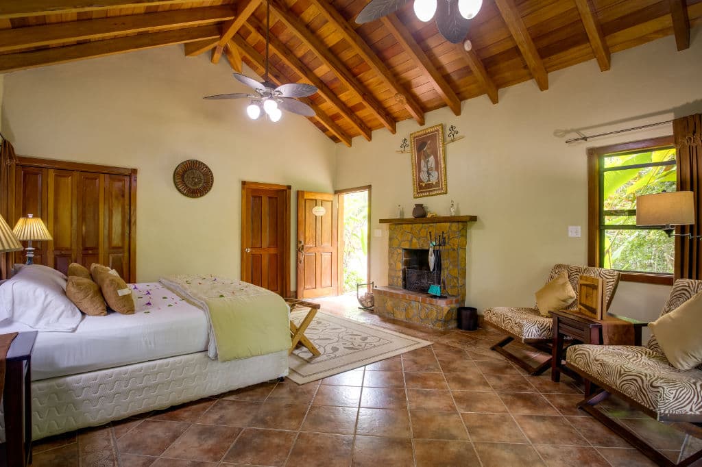 A cozy bedroom with a high wooden ceiling, featuring a bed, seating area, and a stone fireplace.