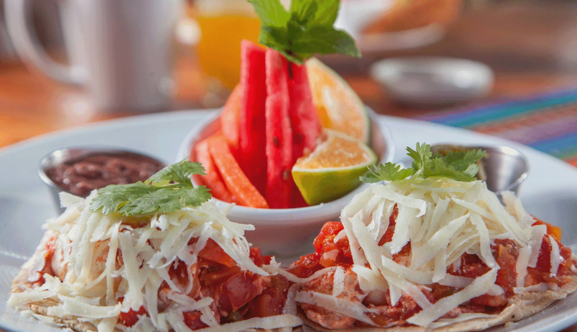 A plate of two tacos topped with shredded cheese and cilantro, accompanied by a fruit platter of watermelon and orange slices, along with a side of black beans.